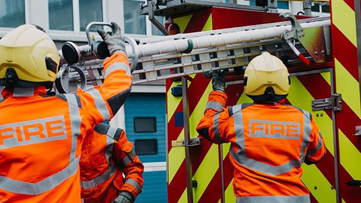 Two firefighters in orange jackets marked FIRE lift a ladder down from a fire engine