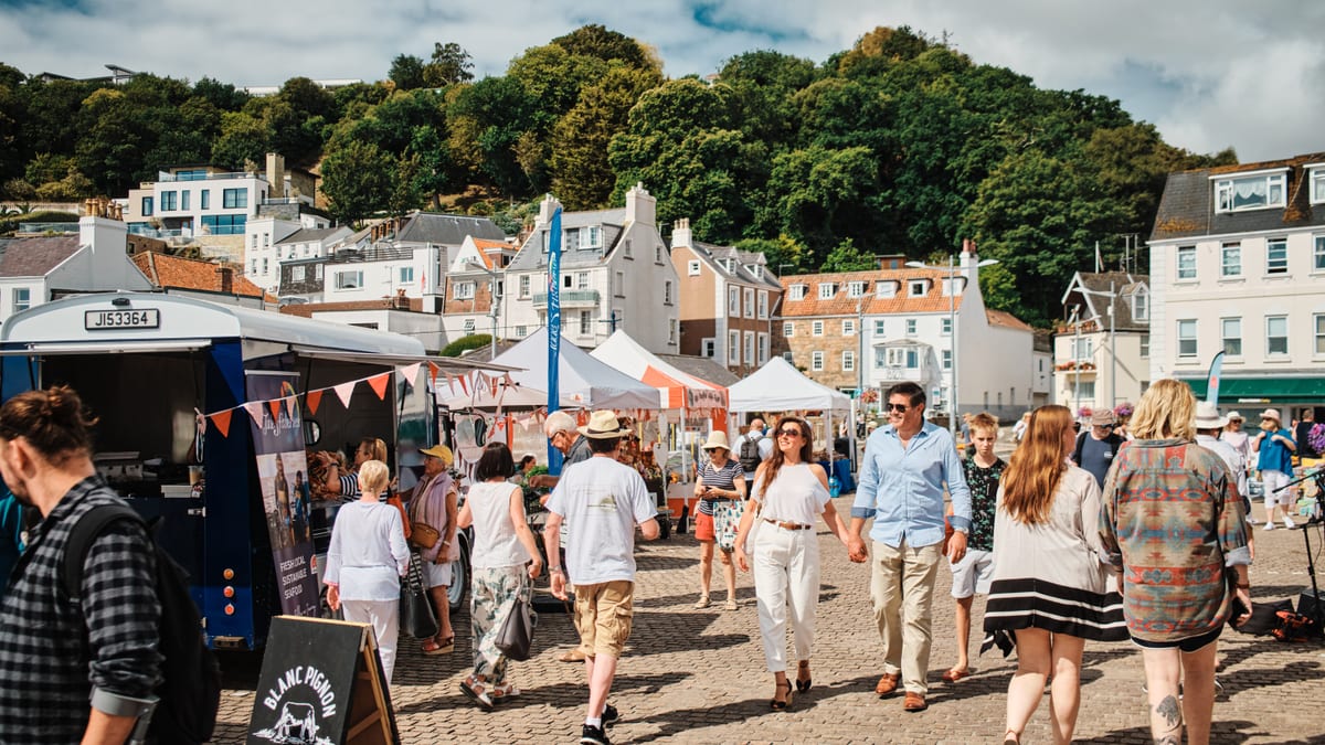 People walking through a sunny harbourside market in Jersey, with stalls, a food van and houses on the wooded hill behind
