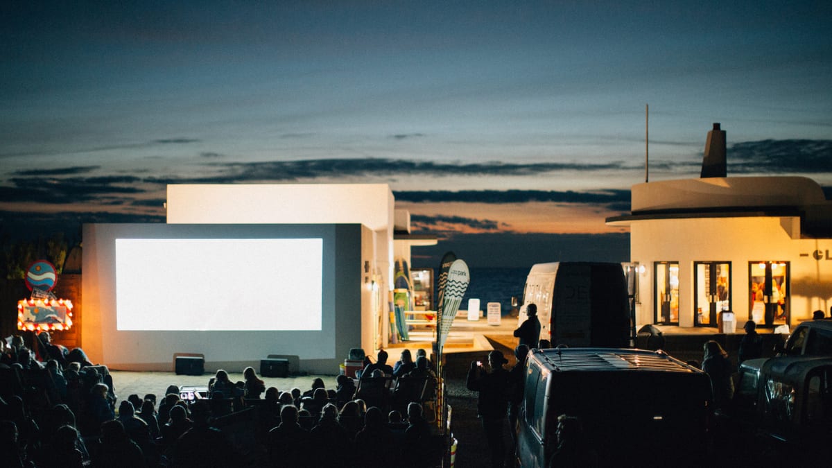 An audience seated at an outdoor cinema at dusk beside a beach kiosk, with the sea behind